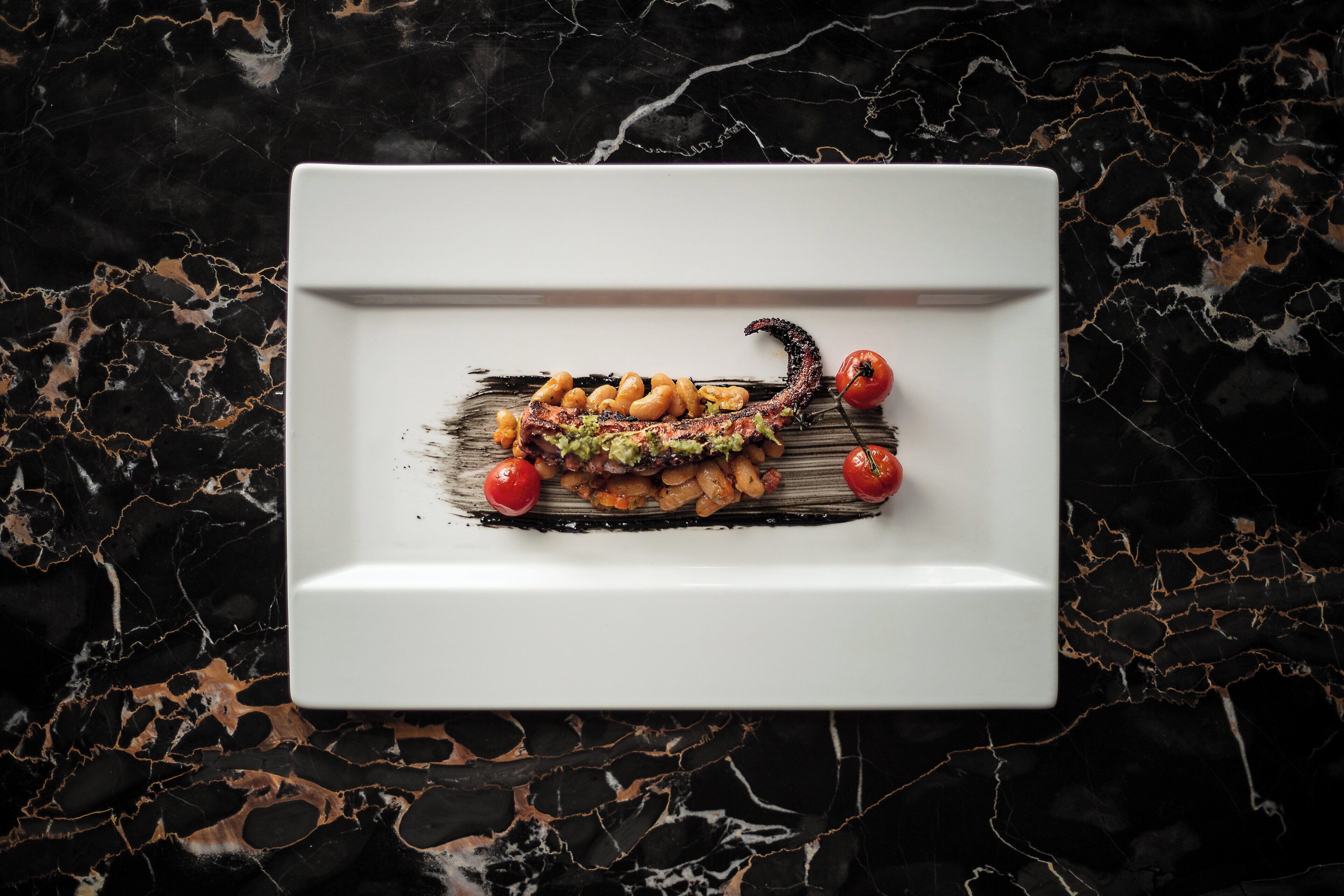 Un plato blanco cuadrado con una porción de comida, que incluye carne, verduras y frijoles, sobre una superficie de mármol oscuro.
