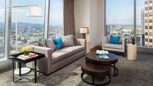 Moderni hotellihuone, jossa on suuri ikkuna, josta avautuu näkymä kaupunkiin, sohva, nojatuoli, pieni pöytä ja lamppu.