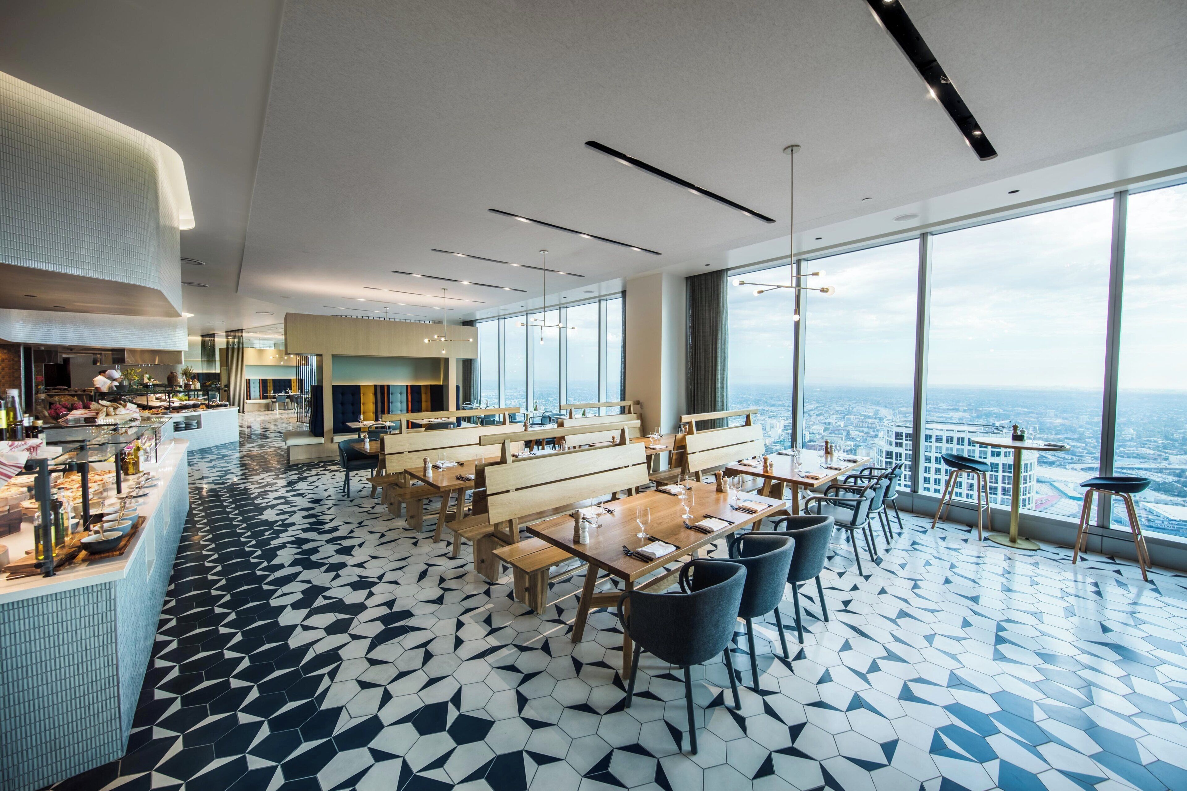Ein modernes Restaurant mit hoher Decke, einem geometrisch gemusterten Boden und großen Fenstern mit Blick auf die Stadt.