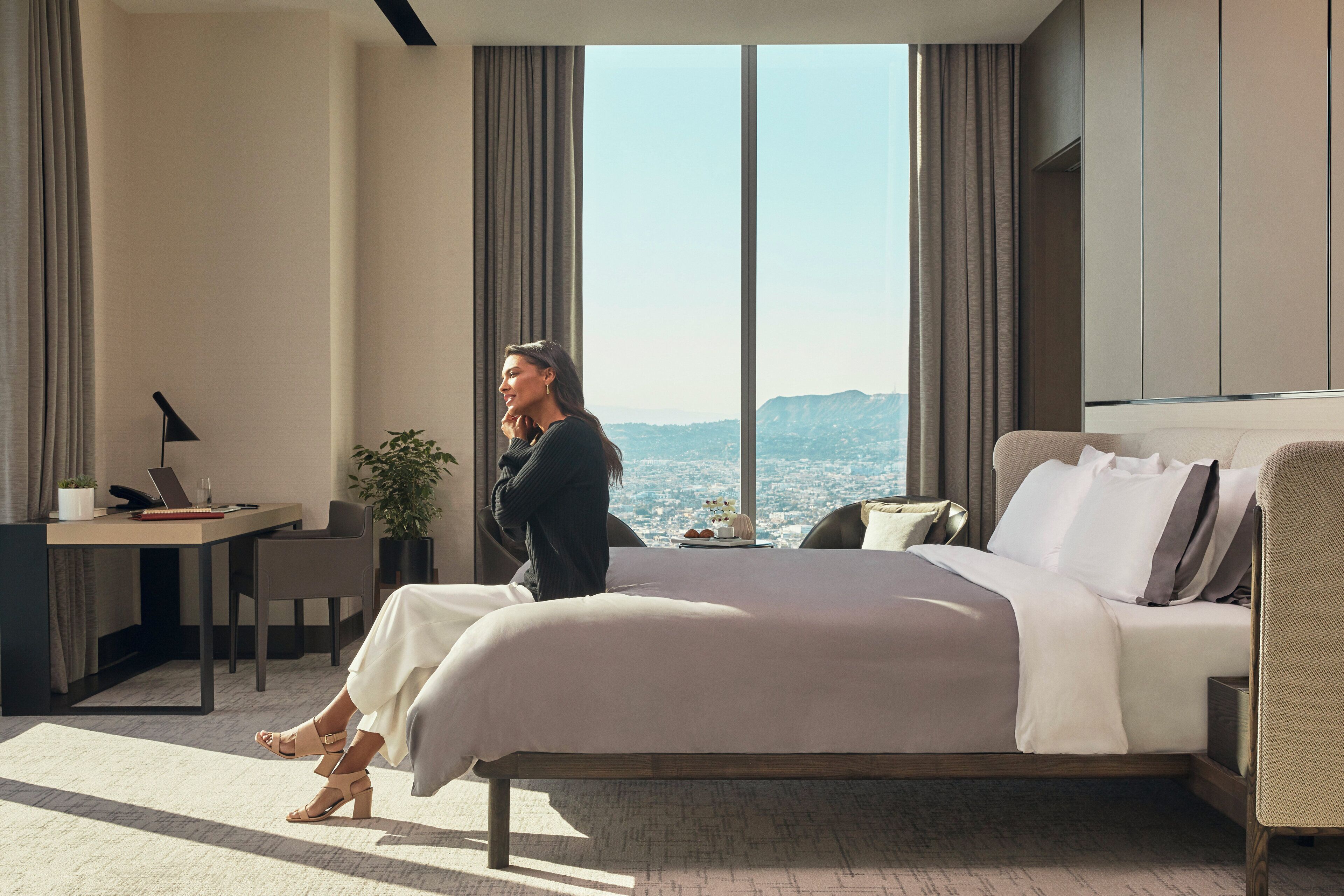 A modern hotel room with a large bed, a desk, and a view of the cityscape.