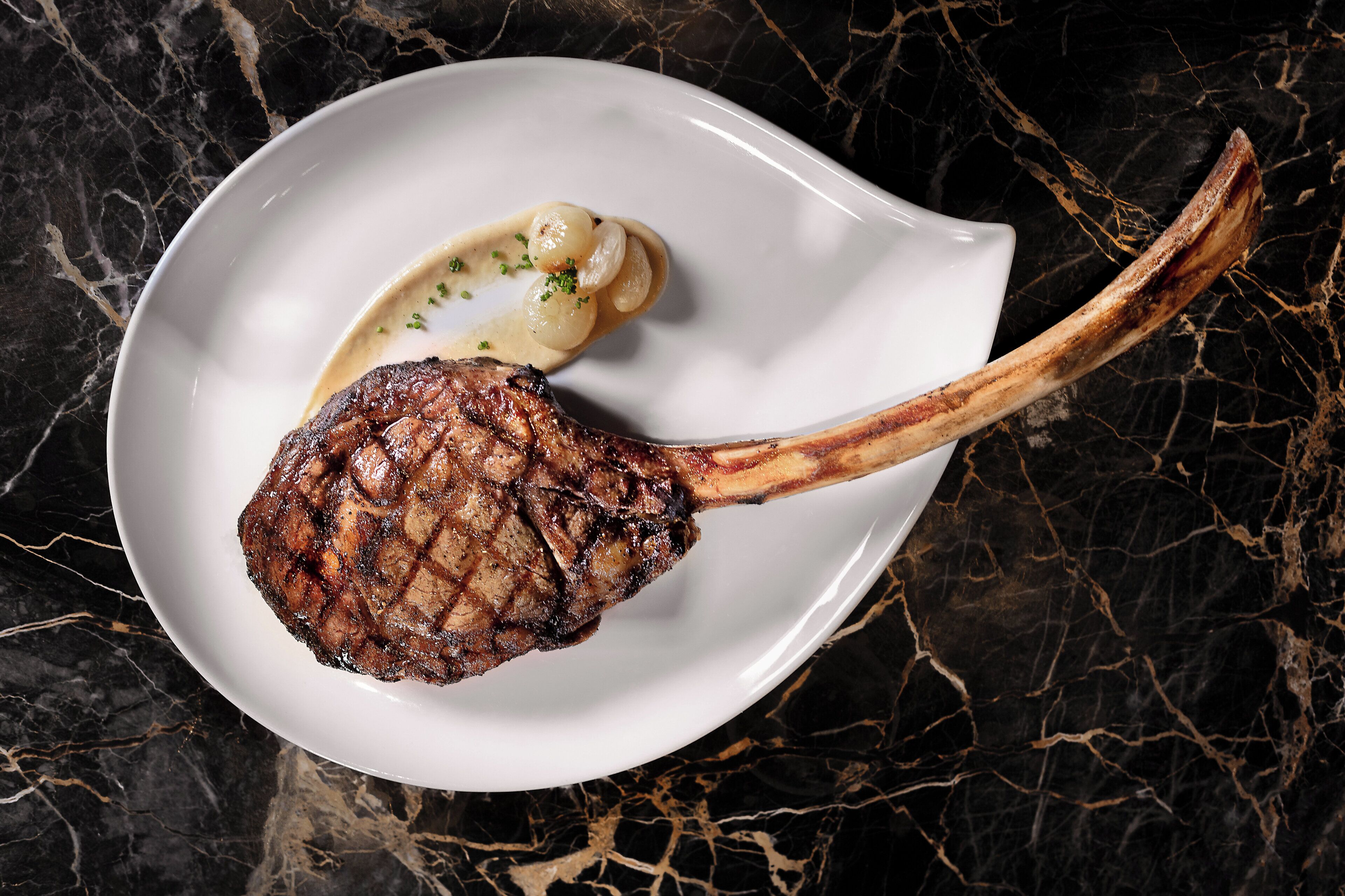 A steak with a bone on a white plate, set against a dark marble background.
