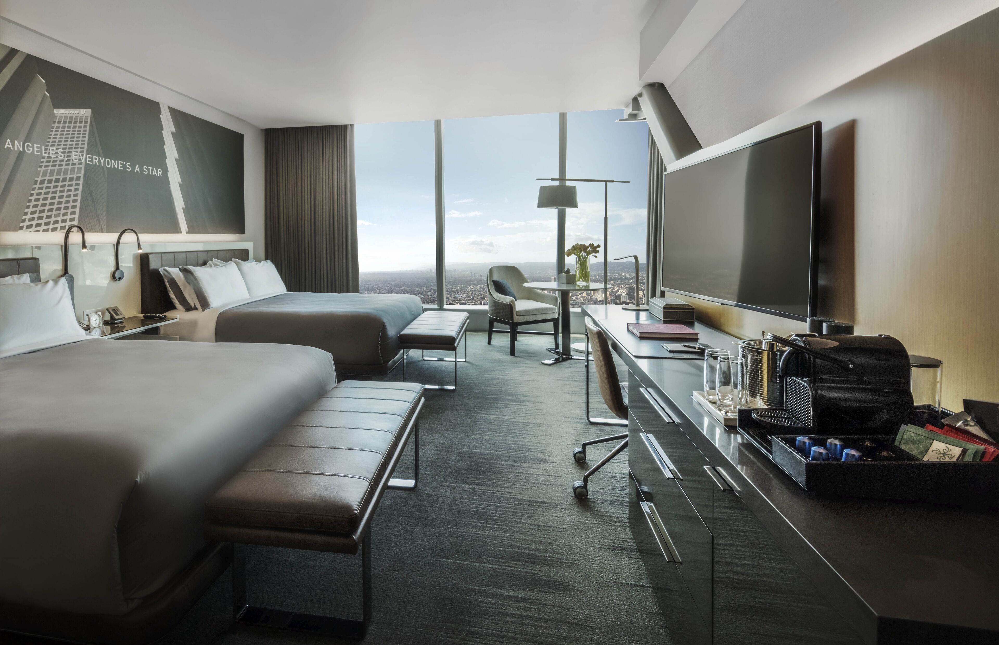 Quarto de hotel moderno com uma cama grande, uma escrivaninha com televisão, uma cadeira e vista para a cidade.