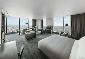 Ein modernes Hotelzimmer mit einem großen Bett, einer Couch, einem Schreibtisch und einem Fernseher. Das Zimmer hat große Fenster mit Blick auf die Stadt.