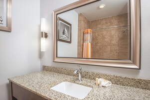 Une salle de bain dotée d’un comptoir en granit, d’un lavabo blanc, d’un miroir encadré et d’une douche avec une porte en verre.