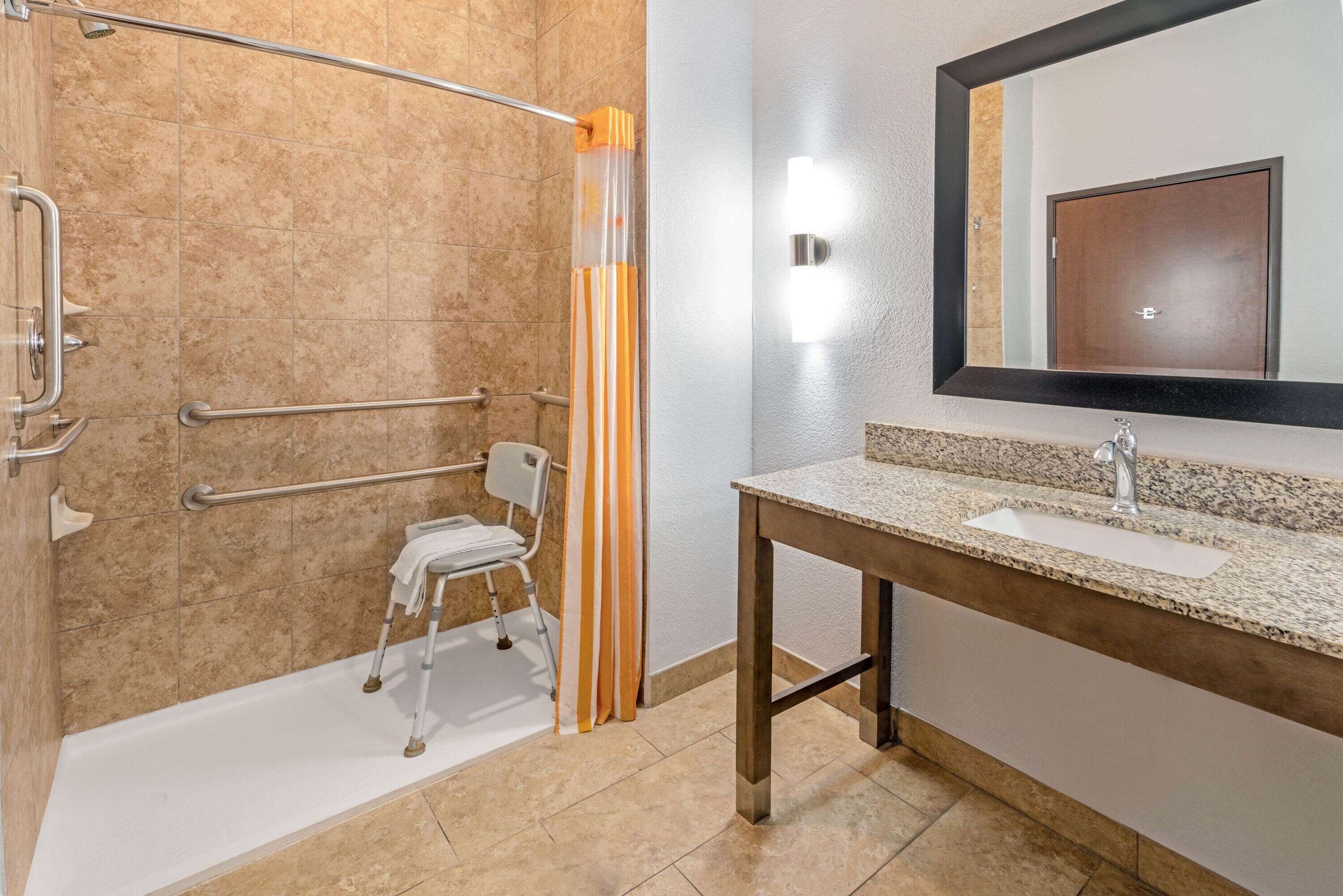 Une salle de bain moderne équipée d’une douche, de barres d’appui, d’un siège accessible aux personnes en fauteuil roulant, d’un lavabo et d’un grand miroir.
