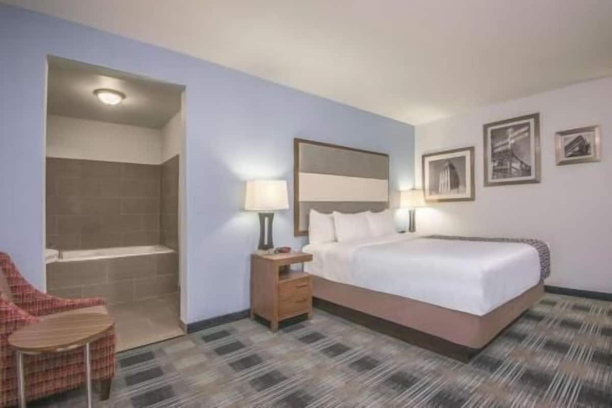 A hotel room with a bed, a chair, a nightstand, a lamp, a shower, and a wall with framed pictures.