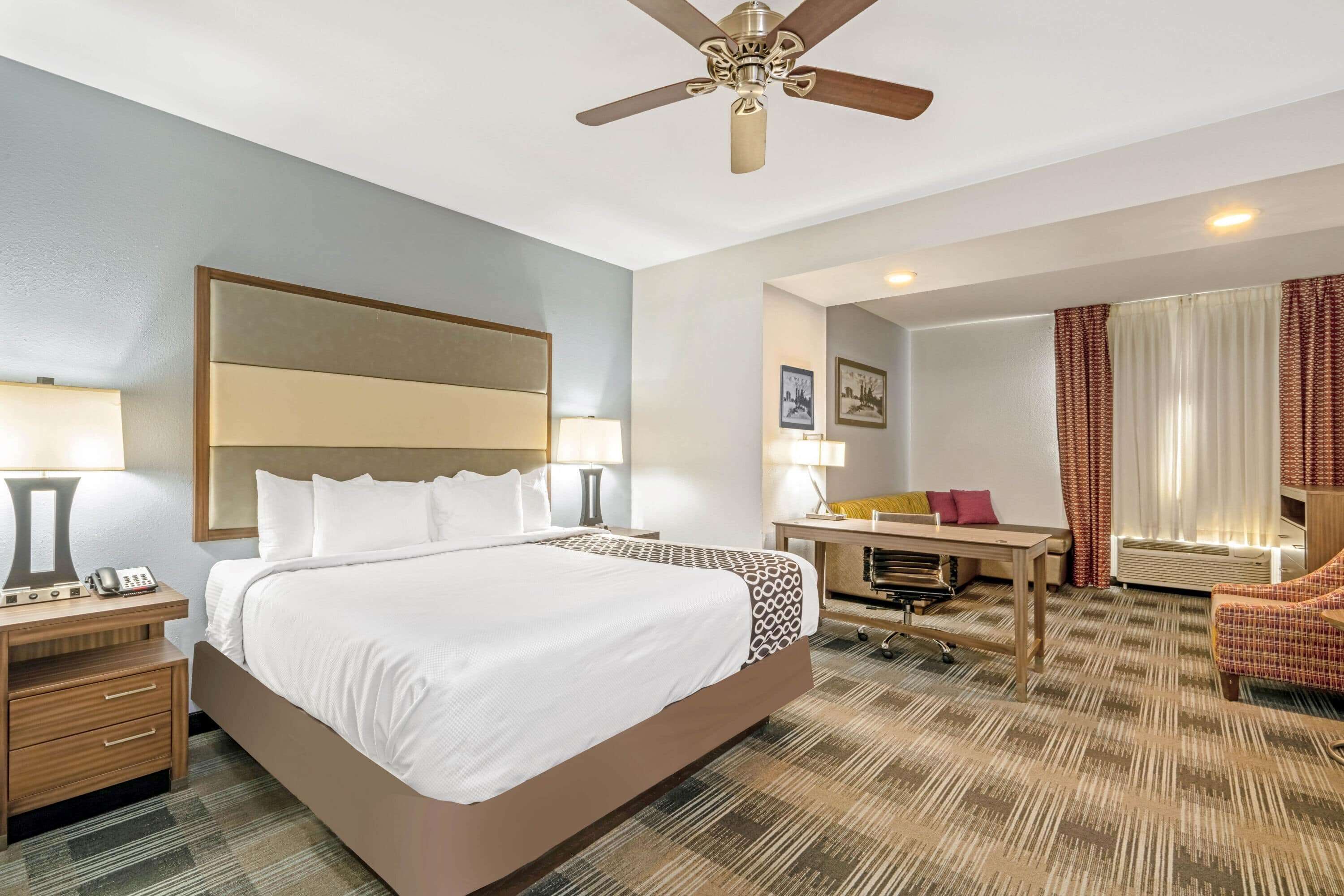 A hotel room with a large bed, a desk, a chair, a ceiling fan, and a window with curtains.