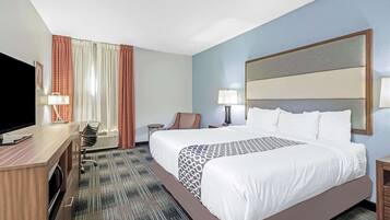 A hotel room with a large bed, a desk with a flat-screen TV, a chair, a lamp, and a window with curtains.
