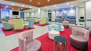 A hotel lobby with a red carpet, various seating options including armchairs and ottomans, a television mounted on the wall, and a large window with a view of a building.