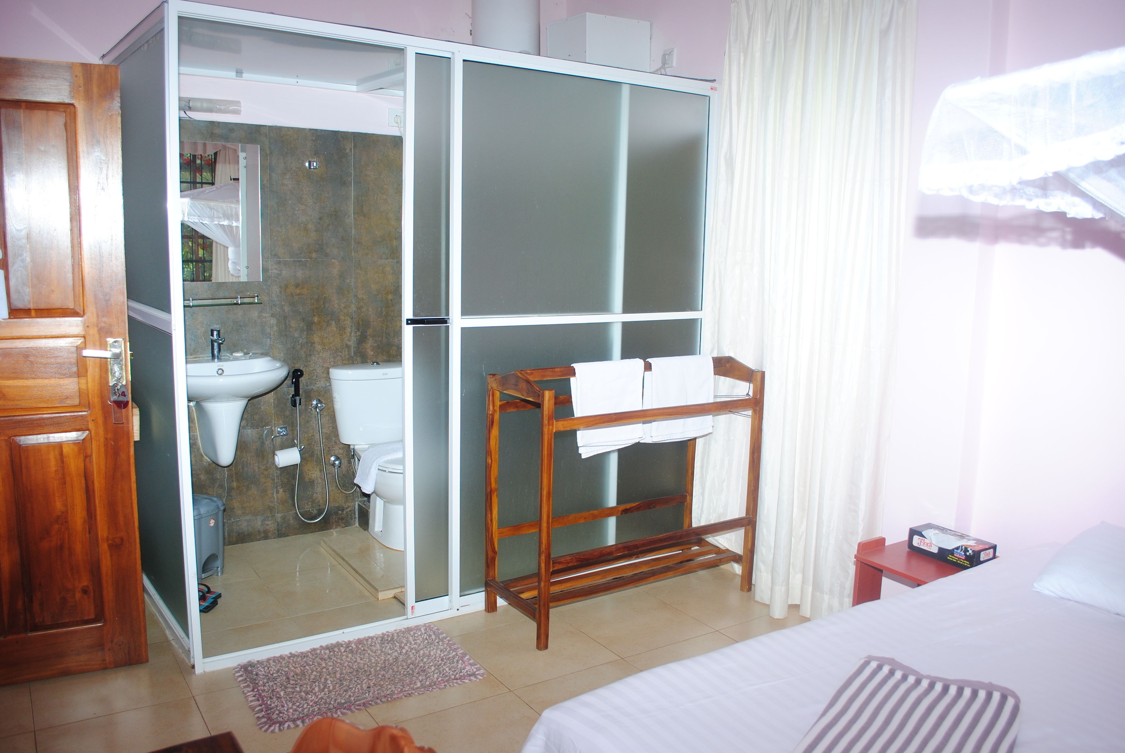 Habitación de hotel con un armario de madera, baño con lavabo y espejo, tendedero de madera, cama con almohada a rayas y una ventana con cortinas.