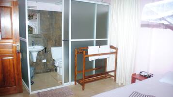 Habitación de hotel con un armario de madera, baño con lavabo y espejo, tendedero de madera, cama con almohada a rayas y una ventana con cortinas.