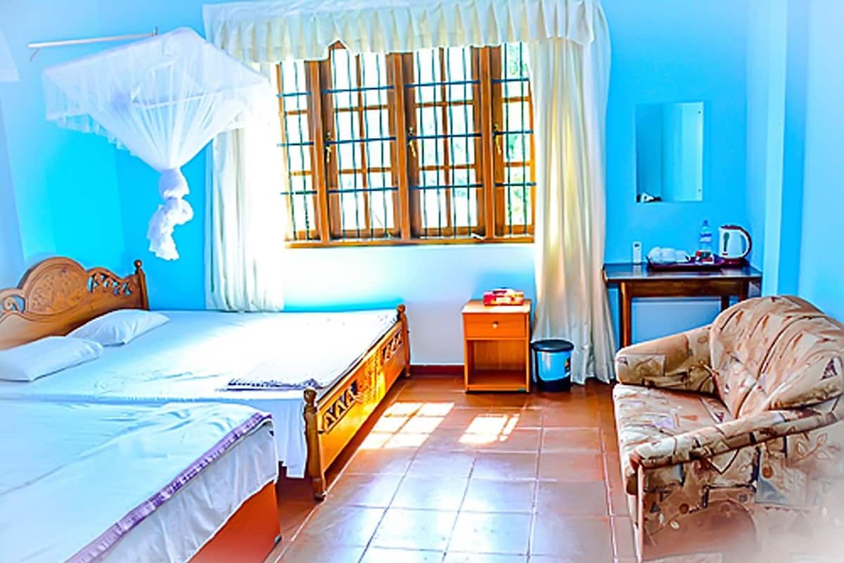 A bedroom with a bed, a bedside table, a nightstand, a chair, a window with curtains, and a small table with a kettle.