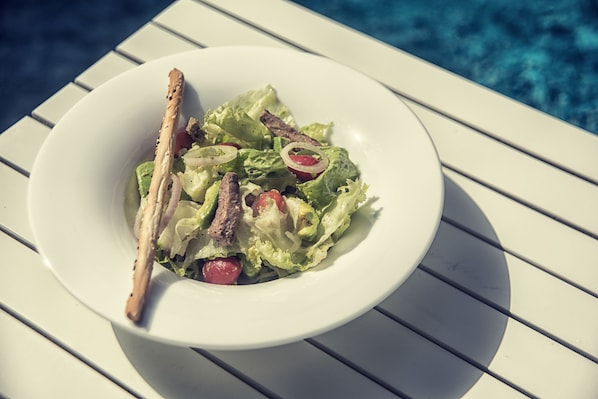 A white bowl of salad with lettuce, tomatoes, and a piece of grilled meat, placed on a white table by a pool.