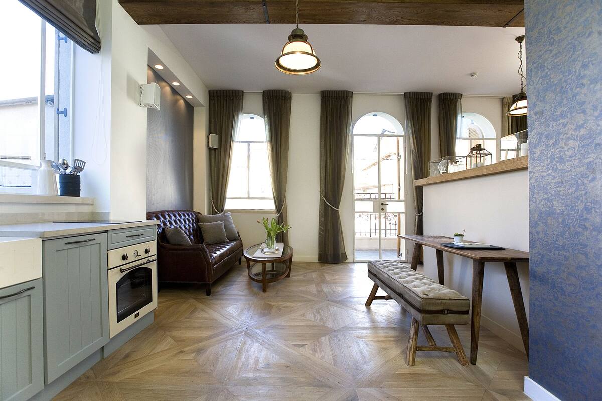 A modern kitchen with a dining area, wooden flooring, and a large window with curtains.