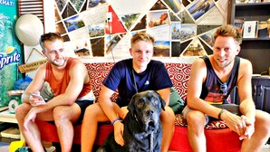 Three people sitting on a couch with a large black dog in a room with a wall decorated with photos and notes. - Danang Backpackers Hostel (Da Nang)