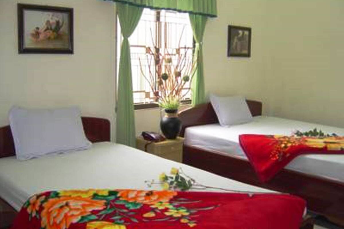 A hotel room with two single beds, a window with green curtains, and a vase with flowers on the nightstand.