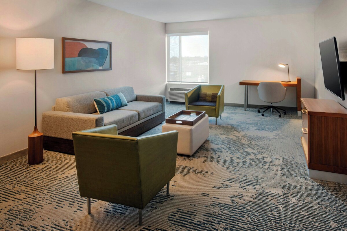 A hotel room with a sofa, armchair, desk, TV, and a painting on the wall.