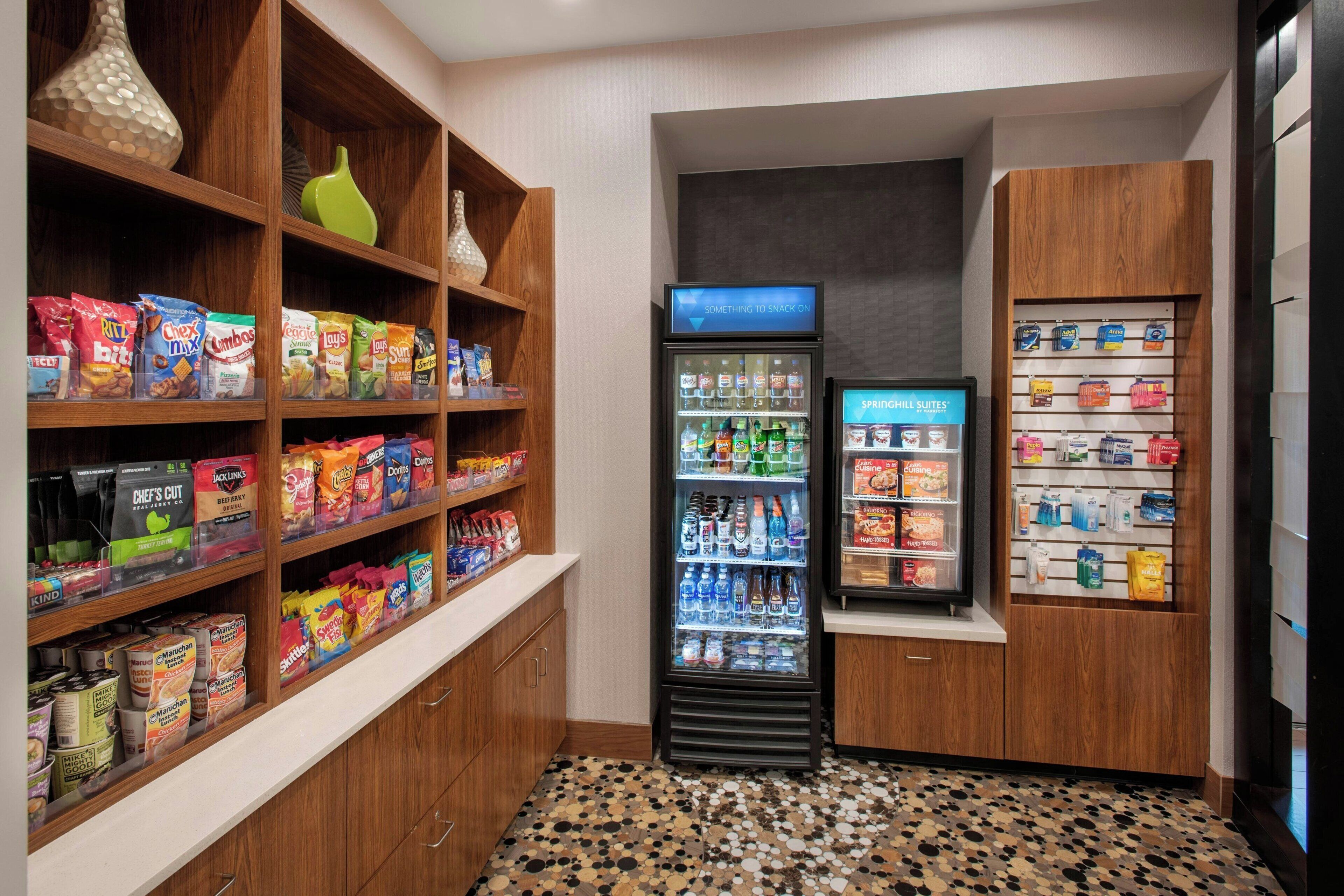 A hotel pantry with shelves stocked with snacks, a refrigerated section, and a display for bottled water.