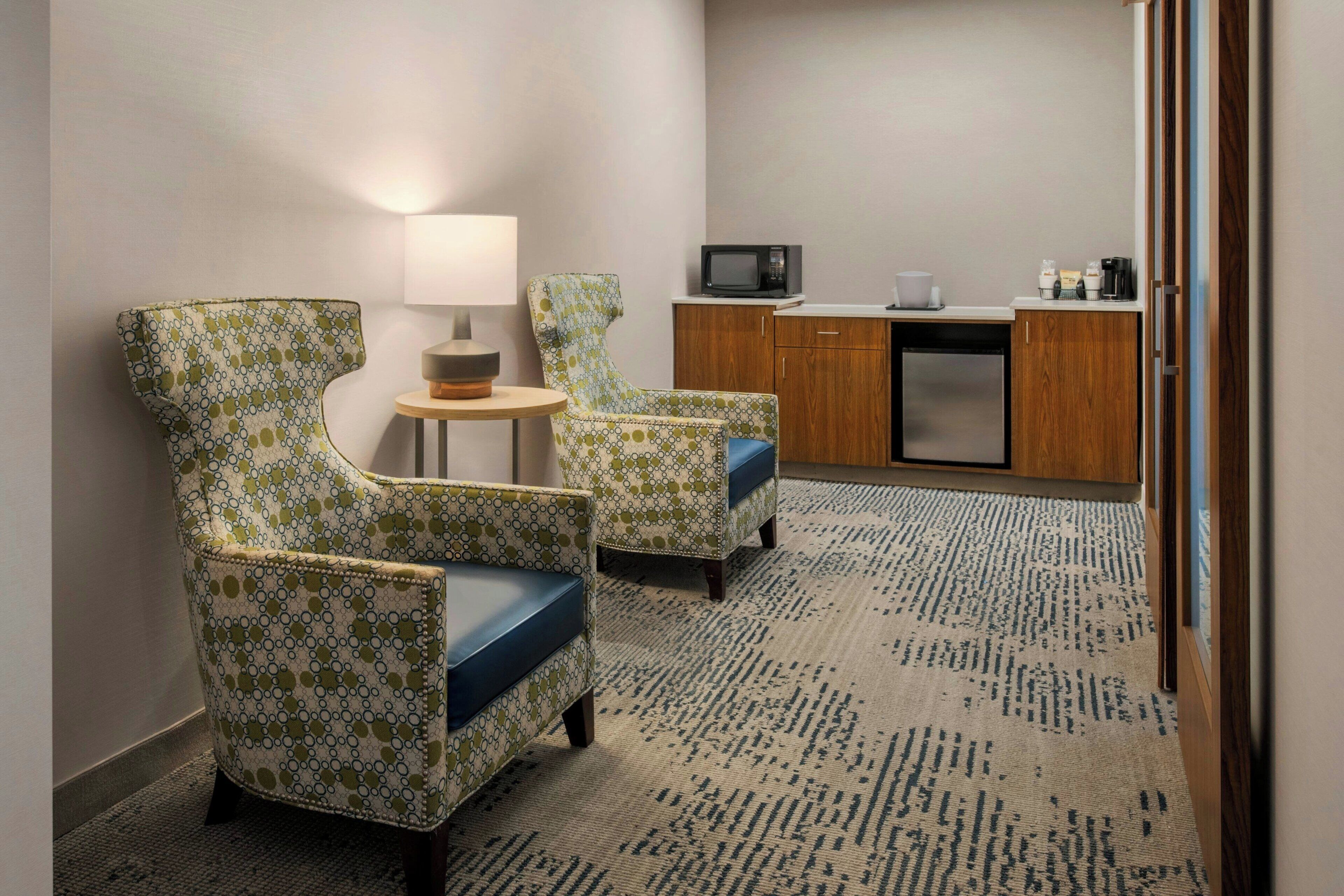 A hotel room with two patterned armchairs, a small round table with a lamp, a kitchenette with a microwave, and a patterned carpet.