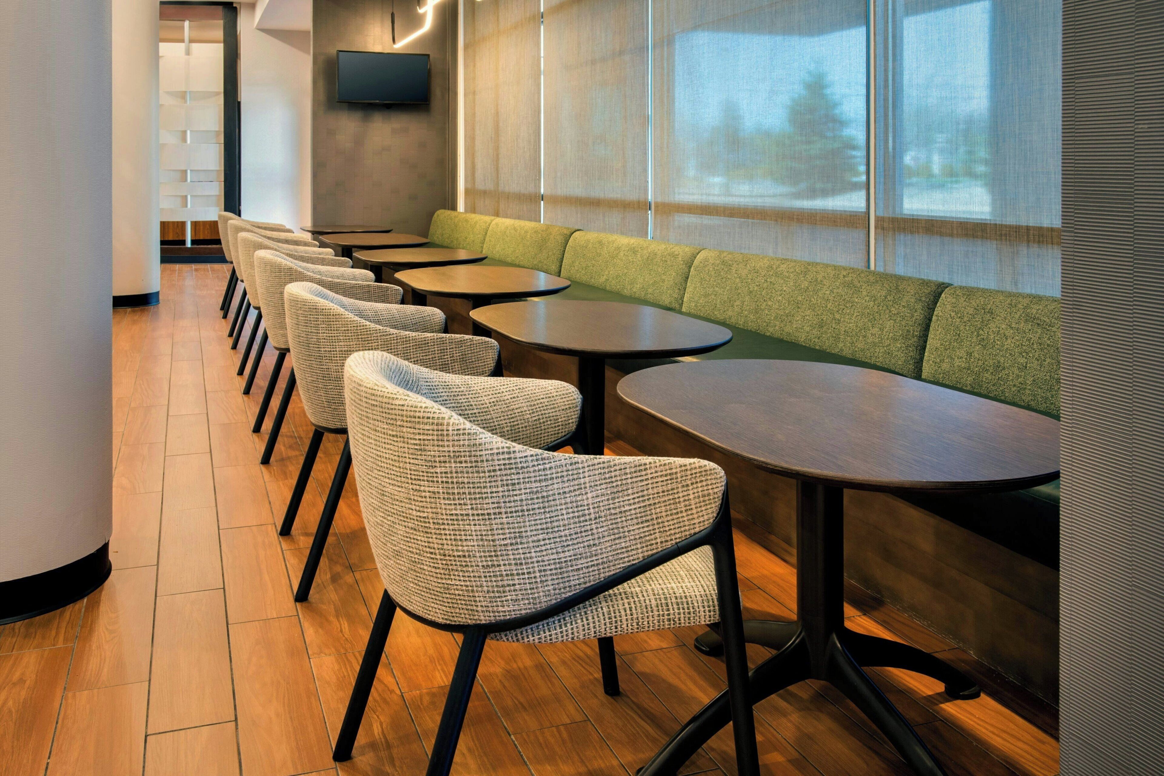 A modern interior with a row of wicker chairs and tables, a green cushioned bench, and a large window with a view.