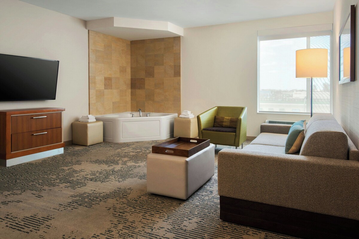 A hotel room with a flat-screen TV, a bathtub, a green armchair, a gray sofa, and a large window with a view.
