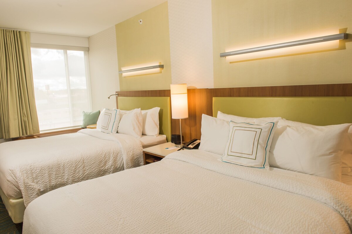 A hotel room with two beds, a nightstand, a lamp, and a window with curtains.