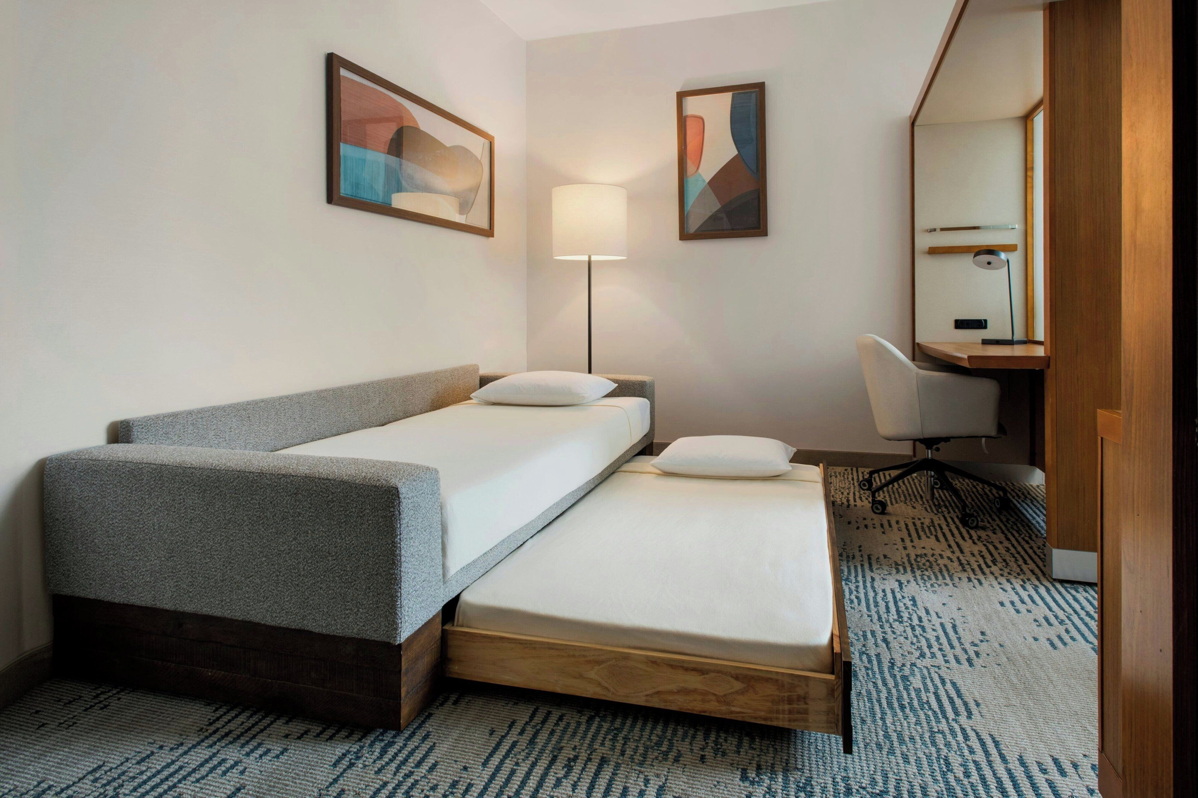 A hotel room with a bed, a desk, a chair, a lamp, and two abstract paintings on the wall.