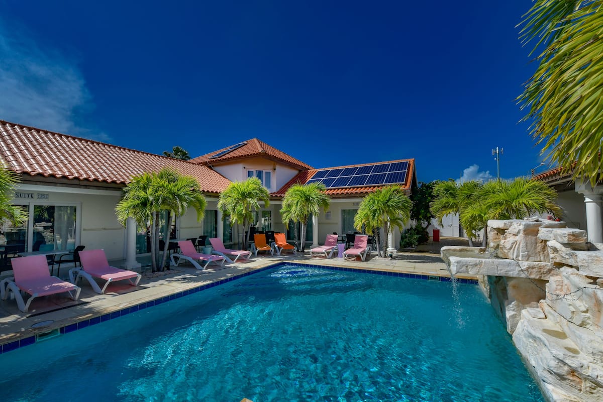 A swimming pool with lounge chairs, a building with a red-tiled roof, and solar panels.