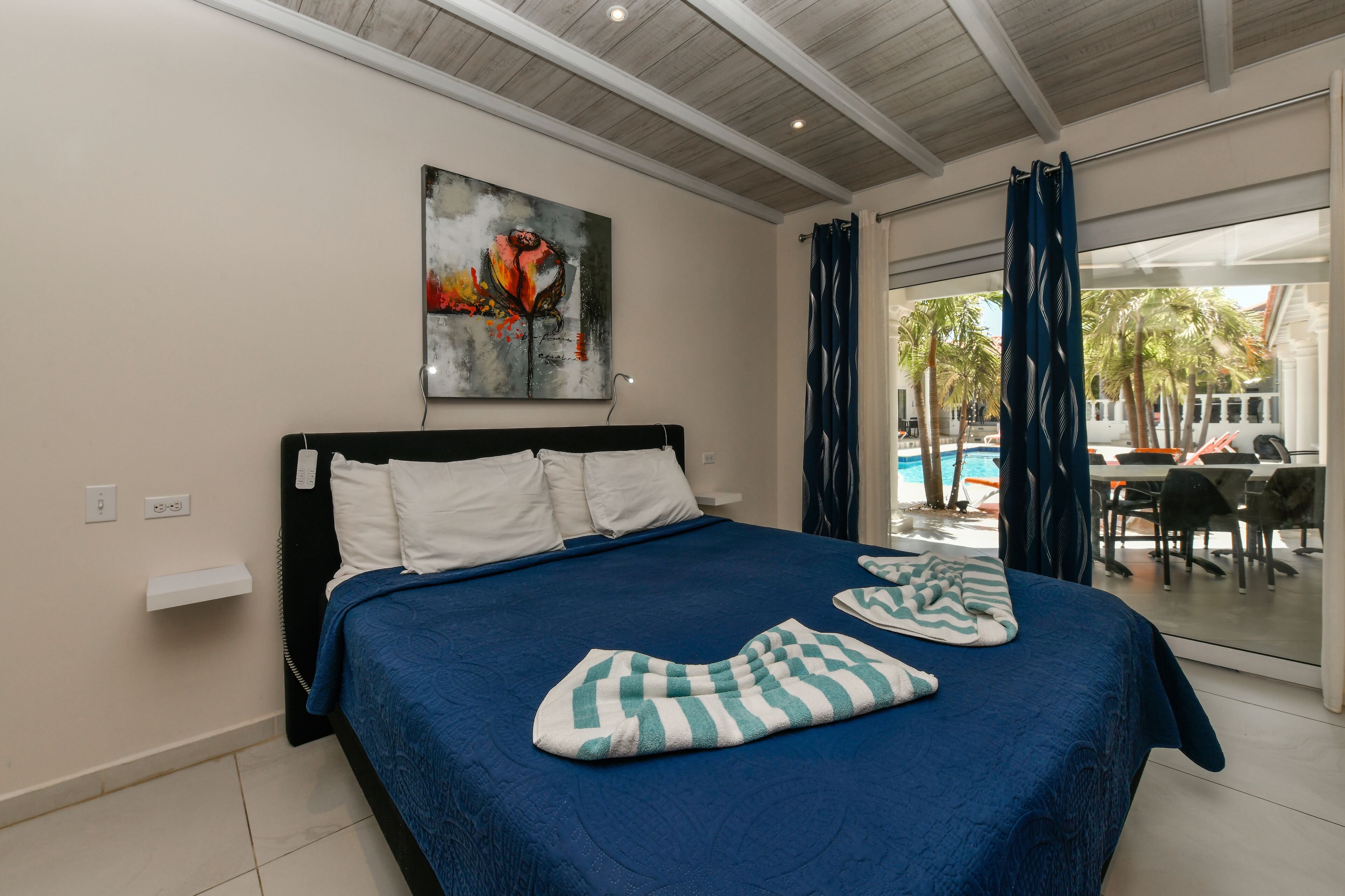 A hotel room with a large bed, a painting on the wall, and a view of the pool and outdoor seating area.