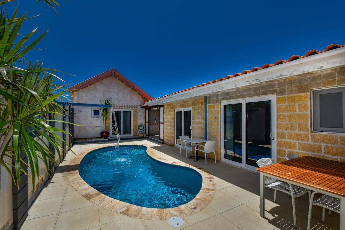 A swimming pool with a curved edge, a patio area with a table and chairs, and a house with a tiled roof and stone walls.