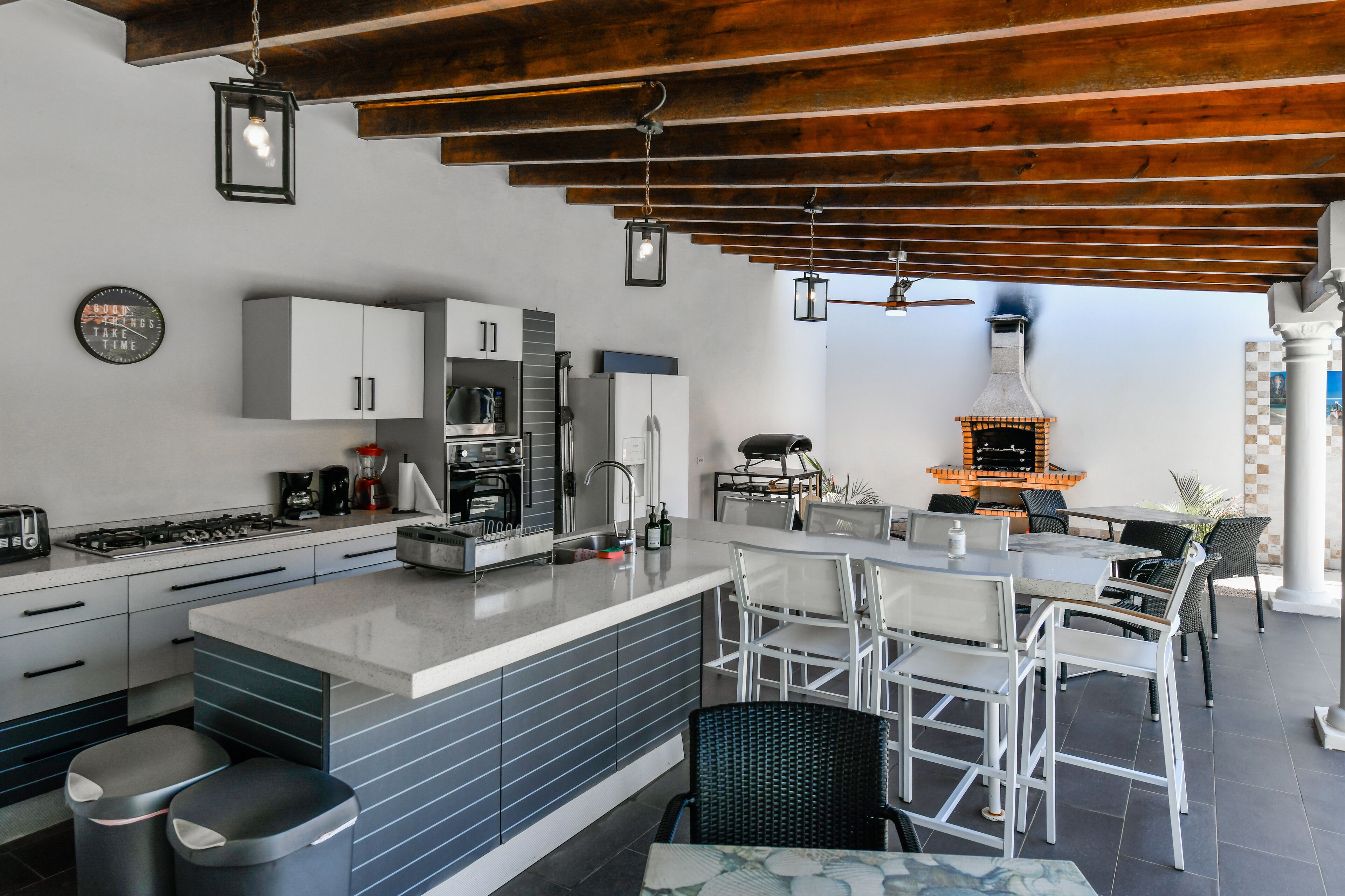 A modern kitchen with a central island, white cabinetry, and a built-in stove. There is a dining area with white chairs and a table, and a wood-fired oven in the background.