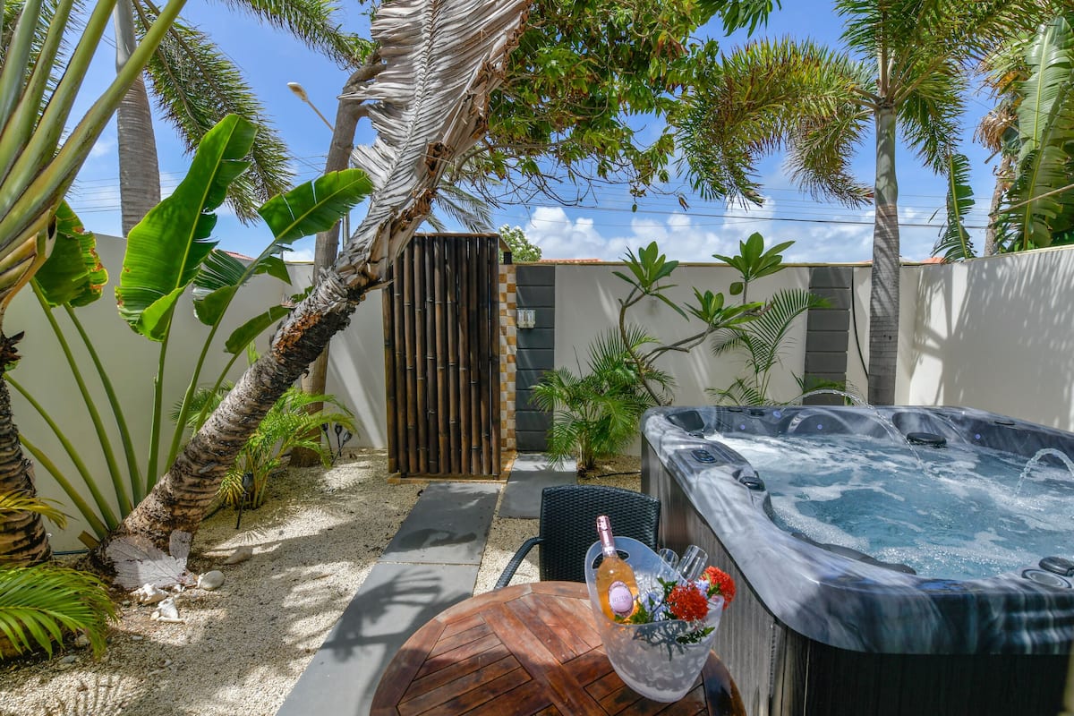 A hot tub with a bottle of champagne and flowers, surrounded by tropical plants and a bamboo fence.
