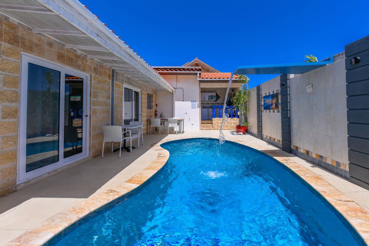 A swimming pool with a tiled edge, a covered patio area with outdoor furniture, and a building with a tiled roof and a door with a glass panel.