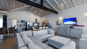 Un amplio salón con un sofá modular grande, una mesa de comedor con sillas, una cocina al fondo y un televisor de pantalla plana montado en la pared.