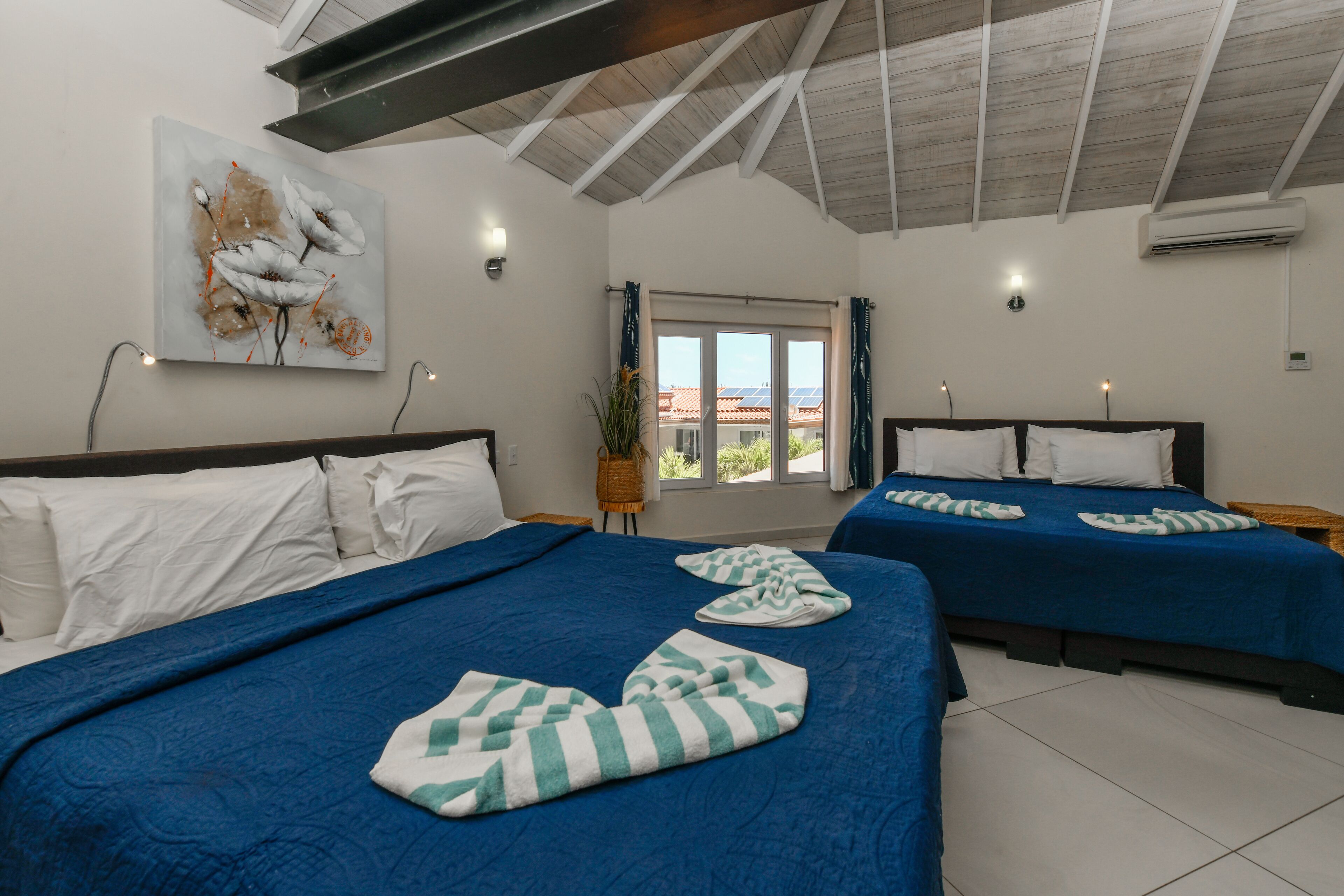 Habitación de hotel con dos camas, cada una con ropa de cama azul y almohadas blancas. En la pared sobre las camas hay un cuadro, una ventana con cortinas y un equipo de aire acondicionado.