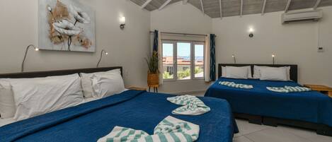 Habitación de hotel con dos camas, cada una con ropa de cama azul y almohadas blancas. En la pared sobre las camas hay un cuadro, una ventana con cortinas y un equipo de aire acondicionado.