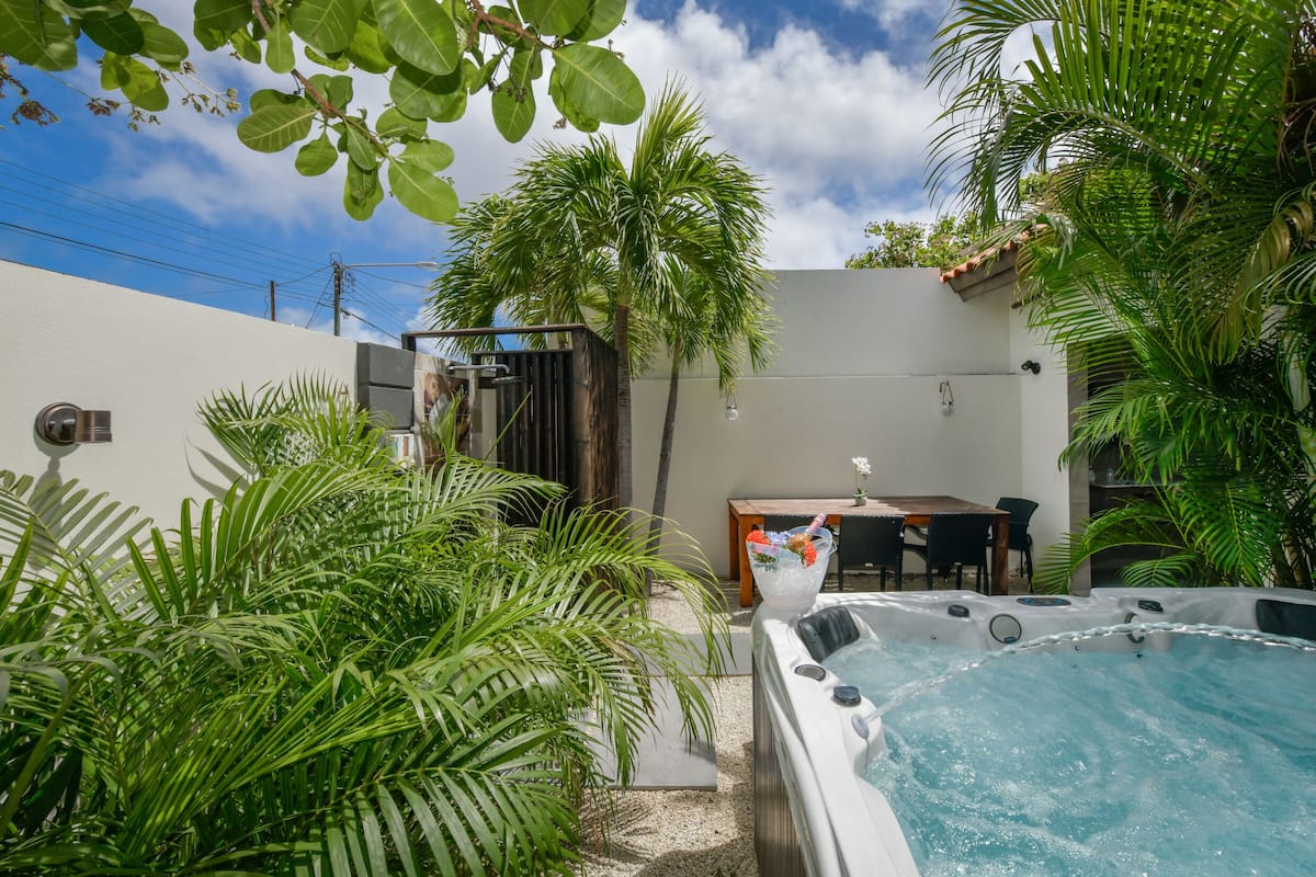 A hot tub surrounded by lush greenery, a dining table, and a white wall.