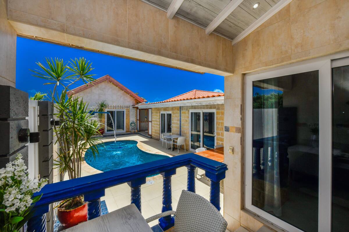 A covered outdoor area with a swimming pool, patio furniture, and a view of a house with a red roof.