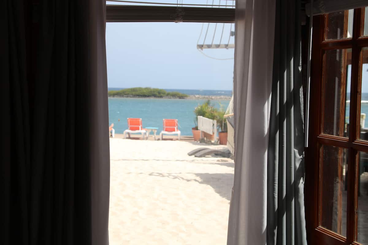 Vista a la playa a través de una ventana con marco de madera, cortinas y una vista del mar, sillas de playa y una isla distante.