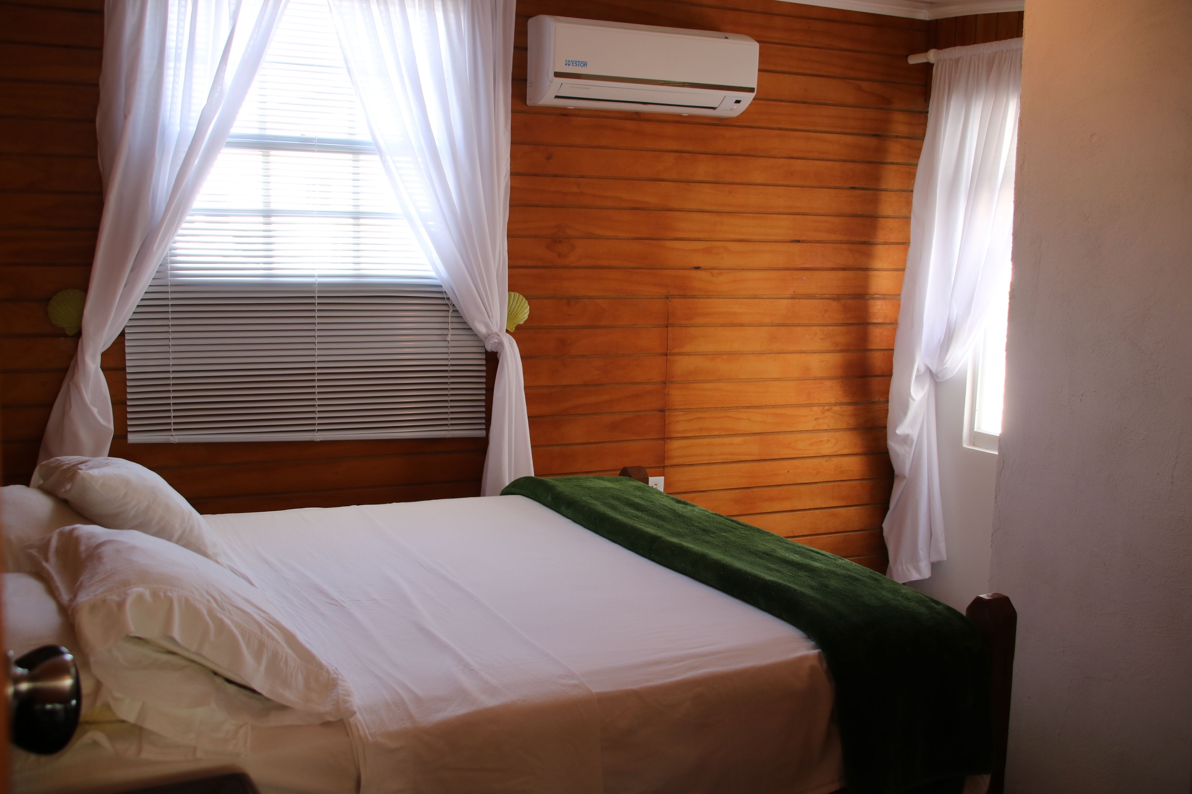 Una cama bien hecha con sábanas blancas, un plaid verde y un cabecero de madera. Hay una ventana con cortinas blancas y un equipo de aire acondicionado montado en la pared.