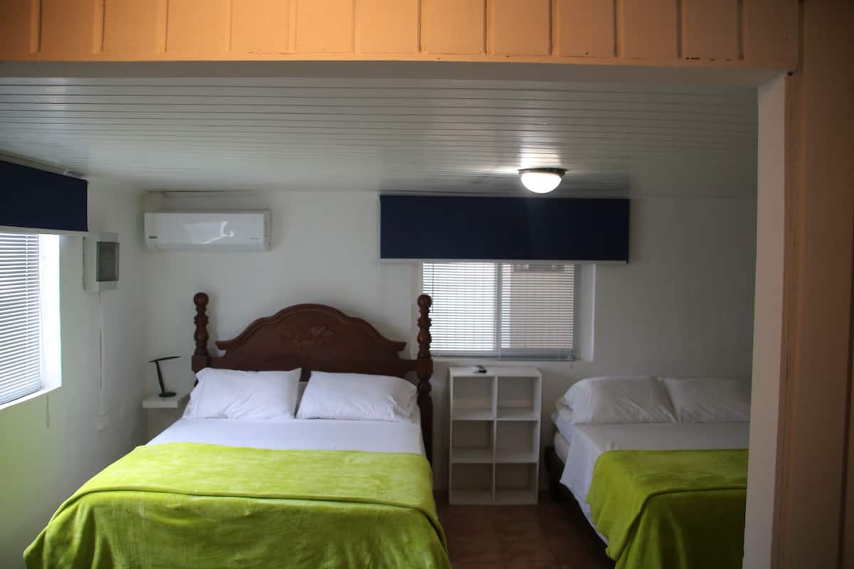 Un dormitorio con dos camas, cabecero de madera, aire acondicionado blanco, un estante y una ventana con persianas.
