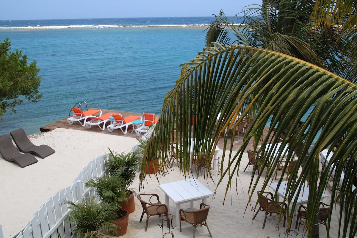 Un patio frente a la playa con sillones, una mesa y sillas, con vista al océano.