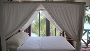 Una cama con ropa blanca y dosel, ventanas amplias y vistas a palmeras.
