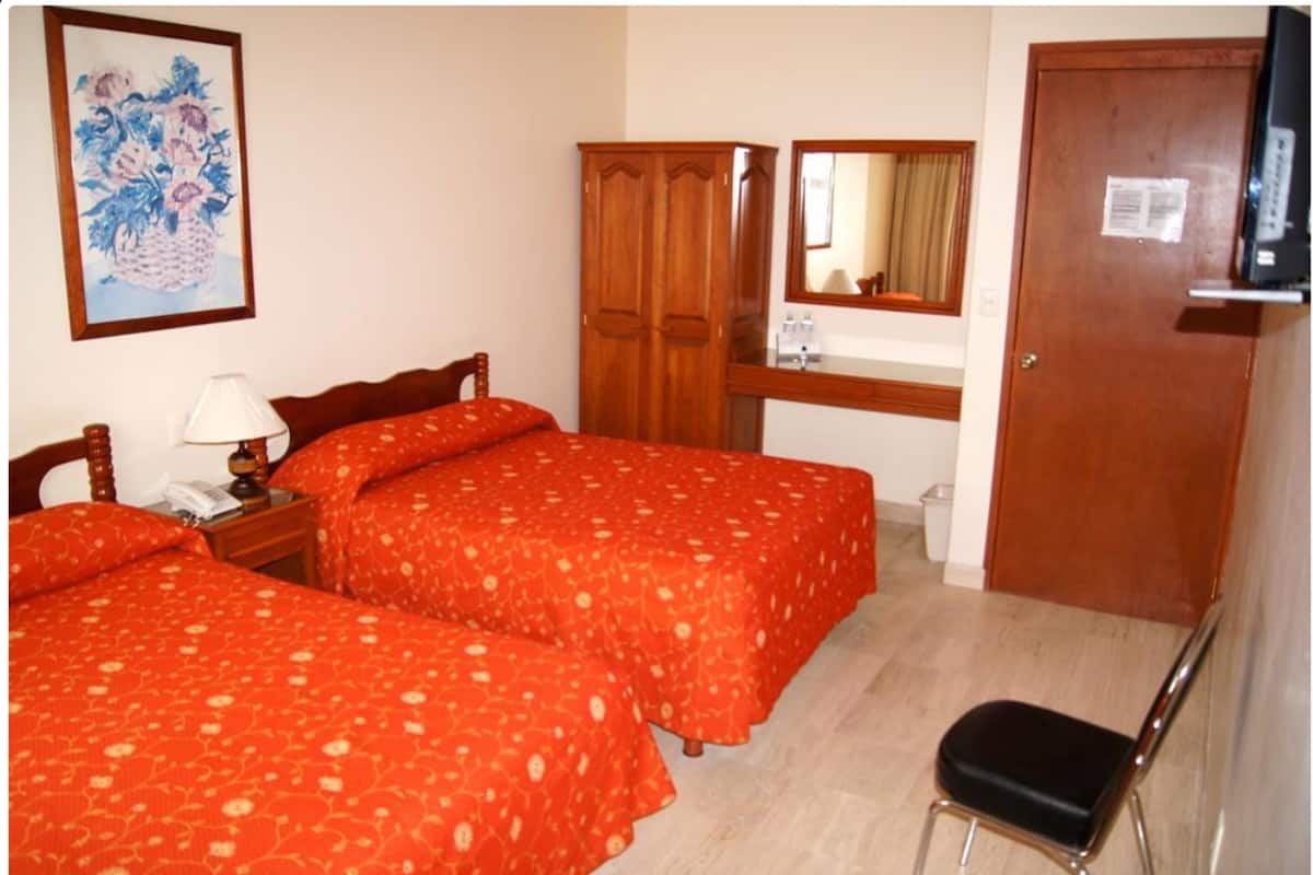 A hotel room with two beds, a wooden wardrobe, a mirror, a desk with a lamp, and a television.