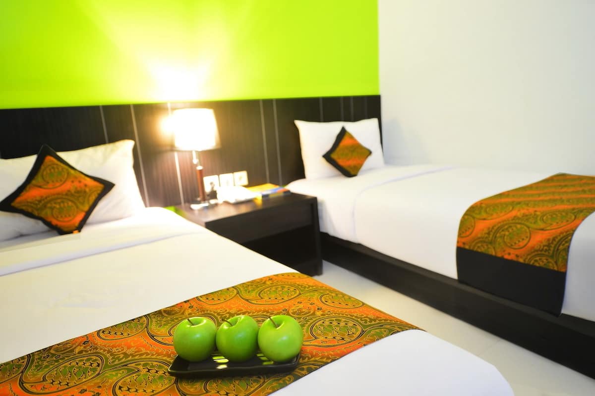 A hotel room with two beds, a nightstand, and a lamp. There are three green apples on a tray on the bed.