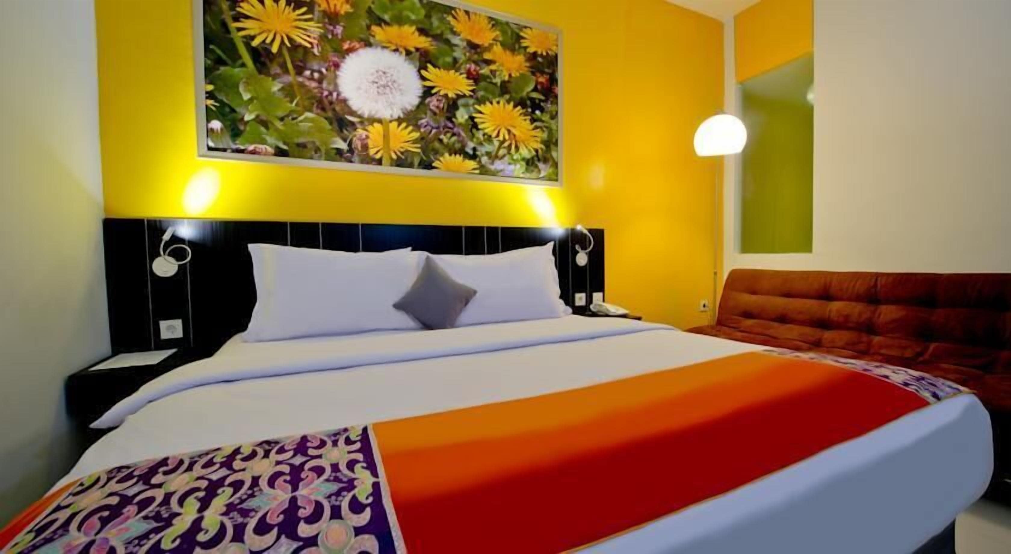 Habitación de hotel con cama, mesita de noche, lámpara y un cuadro de flores en la pared.
