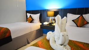 A hotel room with two beds, each with white linens and patterned pillows. There is a nightstand with a lamp and a phone between the beds. A decorative towel arrangement is on the floor.