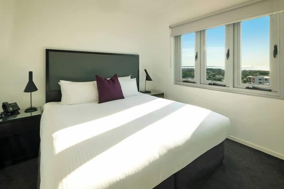 A hotel room with a large bed, a nightstand with a lamp, a window with a city view, and a view of the sky.