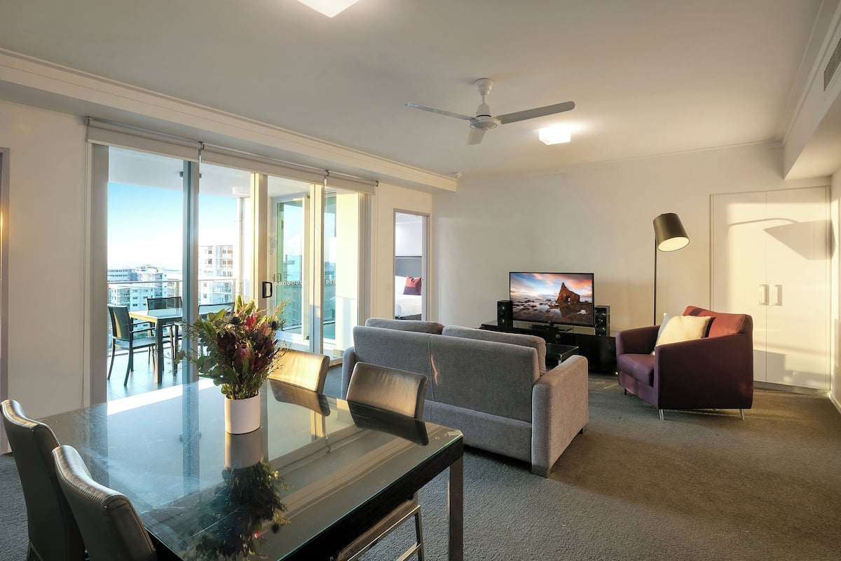 A modern living room with a glass dining table, grey sofas, a purple armchair, a flat-screen TV, and a ceiling fan.