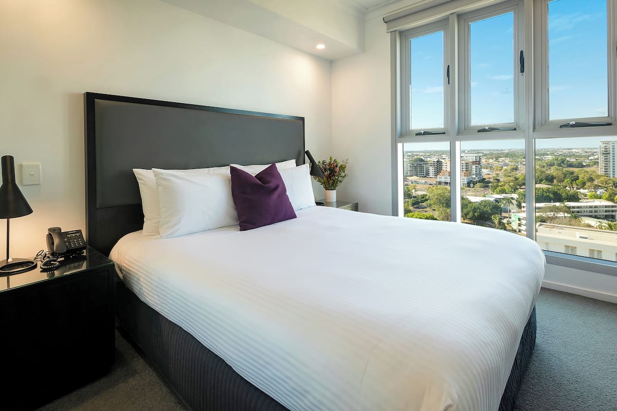 A modern hotel room with a large bed, a nightstand with a phone, and a view of the city through large windows.